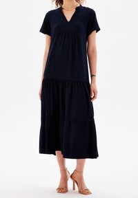 Kvinna iklädd en svart kortärmad midi-klänning med flera lager och V-ringning samt nakenfärgade högklackade sandaler.