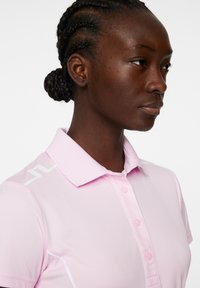 Ljusrosa pikétröja med knäppt krage, med vit accent och logotyp. Slät textur och åtsittande design synlig.