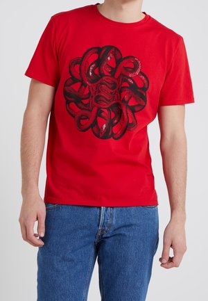 Mężczyzna w czerwonej koszulce z krótkim rękawem z czarnym wzorem splecionych węży ułożonym w okrągły wzór, ubrany w niebieskie dżinsy, stoi na jednolitym tle.