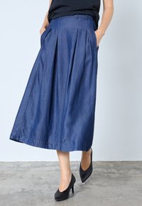 Denim midi svārki ar plisējumiem, dziļi zilā krāsā, ar sānu kabatām un gludu tekstūru. Kopā ar melnām augstpapēžu kurpēm.