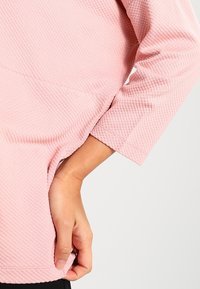 Haut rose clair avec un design texturé, des manches trois-quarts et un ourlet légèrement incurvé. Main visible tenant la couture latérale.