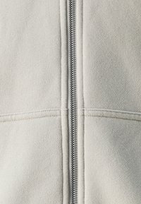 Giacca a zip beige chiaro con zip grigia. Tessuto morbido e cuciture visibili lungo le cuciture. Design semplice.
