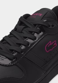 Baskets Lacoste noires avec bordure latérale brillante, logo crocodile rose brodé, lacets noirs et texte de la marque rose sur la languette.