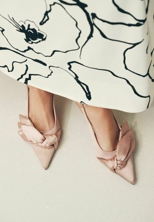 Nohy v bledě růžových saténových lodičkách na špičce s velkou mašlí, částečně zakryté krémovou sukní s výrazným černým abstraktním květinovým potiskem.