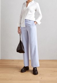 Camicia bianca con bottoni, pantaloni larghi lavanda, borsa triangolare nera e scarpe piattaforma marroni chunky, su uno sfondo chiaro.