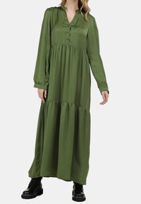 Robe longue verte en tissu léger, semblable à du satin ; dotée d'un plastron à boutons, d'une jupe à volants, de manches longues et de poignets élastiques. Portée avec des bottes noires.