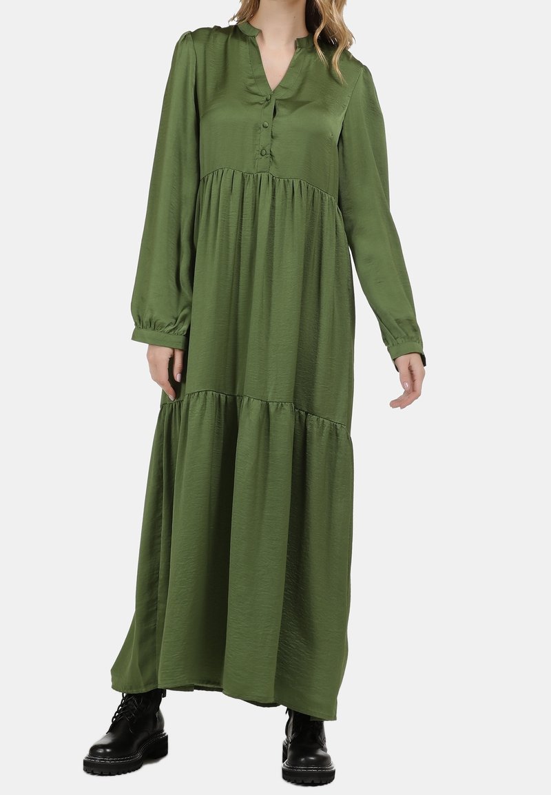 Robe longue verte en tissu léger, semblable à du satin ; dotée d'un plastron à boutons, d'une jupe à volants, de manches longues et de poignets élastiques. Portée avec des bottes noires.