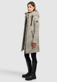 Cappotto piumino grigio chiaro con cappuccio, caratterizzato da un motivo di trapuntatura a zigzag, chiusura con zip e dettagli neri. Abbinato a stivali neri al ginocchio.