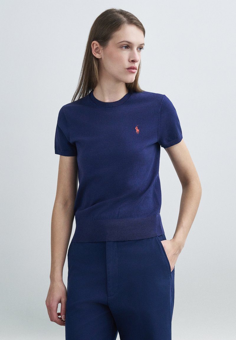 Μπλούζα με κοντά μανίκια και στρογγυλή λαιμόκοψη σε υφή πλεκτού σε ναυτικό μπλε, με μικρό κόκκινο λογότυπο στο αριστερό μέρος του στήθους. Συνδυάζεται με παντελόνι σε ίδιο ναυτικό μπλε χρώμα.