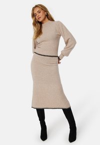 Zweiteiliges Strickoutfit in Beige mit ribbed Struktur. Das Oberteil hat einen runden Halsausschnitt; der Rock weist einen schwarzen Saum am Abschluss auf. Kombiniert mit schwarzen Stiefeln.