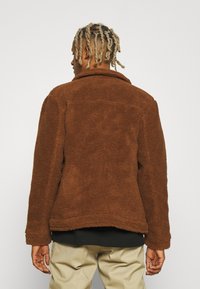 Chaqueta de forro polar marrón con un exterior suave y texturizado, cuello con solapa y un corte holgado, costuras visibles y un dobladillo recto.