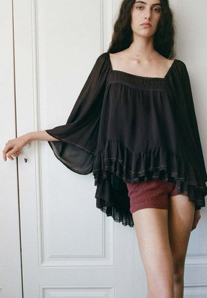 Jeune femme aux longs cheveux foncés porte une blouse noire ample à manches évasées et un short en maille bordeaux, debout devant des portes blanches à panneaux.