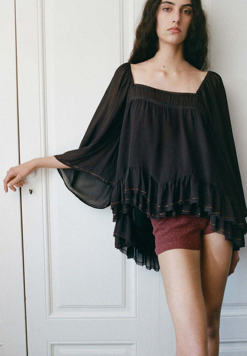 Jeune femme aux longs cheveux foncés porte une blouse noire ample à manches évasées et un short en maille bordeaux, debout devant des portes blanches à panneaux.