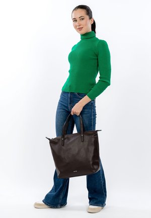 Bolso tote de cuero marrón con doble asa, superficie texturizada y herrajes en tono plateado, sostenido por una persona que lleva un suéter de cuello alto de punto en verde.