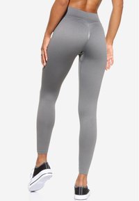 Leggings grigi con vita alta, texture liscia e design aderente, abbinati a scarpe sportive bianche e nere. Dettagli delle cuciture visibili.