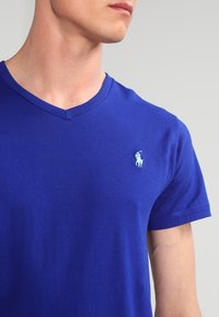 Man i en blå V-ringad T-shirt med en liten vit broderad polo-spelare logotyp på vänster bröst mot en neutral bakgrund.