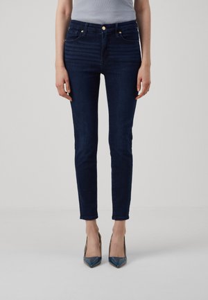Personne portant un jean slim bleu foncé et un haut gris, debout les pieds rapprochés dans des escarpins pointus bleu foncé.