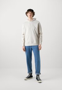 Lys beige hoodie med en forlommet lomme, ribbede manchetter og hætte med snøre. Kombinationen er med oprullede blå denim jeans og sorte højhælede sneakers.
