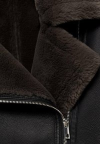 Veste en cuir noire avec col en fourrure douce marron foncé et fermeture éclair argentée visible partiellement ouverte.