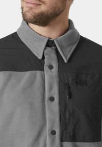 Szary polarowy kurtka z czarnymi akcentami tkaninowymi, zapinana na guziki, z zamkiem błyskawicznym na kieszeni na piersi, z delikatnym logo. Miękka i odporna.