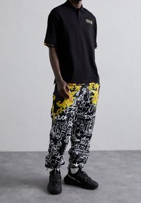 Czarny polo z logo, zestawiony z żółtymi i czarnymi spodniami cargo w wzory ze stylizowanym graffiti oraz luźnym krojem, a także czarnymi trampkami.