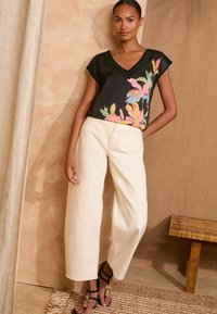 Juodas gėlių marškinėliai trumpomis rankovėmis, su spalvingais lapų raštais. Kreminės spalvos plataus žandikaulio kelnės. Juodi sandalai su dekoratyviniais akcentais.