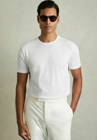 Tricou alb cu mânecă scurtă dintr-un material moale, asociat cu pantaloni cremi, croiți, având un design minimalist și o siluetă ajustată.