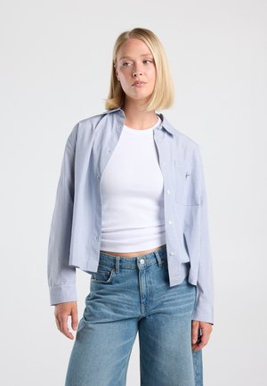 Blondine vrouw draagt een lichtblauw geruit shirt met knopen, open over een wit geribd cropped topje en een blauwe hooggetailleerde spijkerbroek tegen een effen achtergrond.