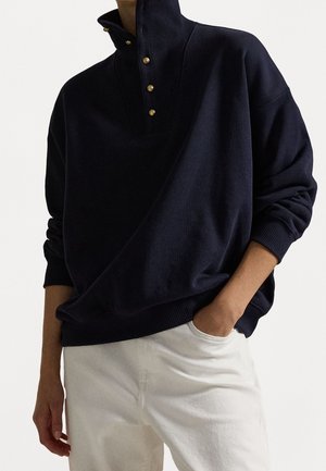 Personne portant un pull bleu marine foncé à col montant avec des boutons dorés, associé à un pantalon blanc, une main dans la poche.