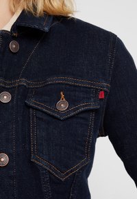Veste en denim foncé avec boutons en cuivre, deux poches poitrine, surpiqûres orange et un col. Présente un accent en étiquette rouge sur la poche.