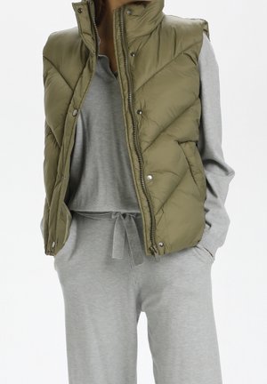 Personne portant un gilet matelassé sans manches vert olive sur un sweat-shirt gris clair et un pantalon à cordon assorti, mains dans les poches, debout devant un fond blanc.