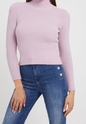 Femme portant un pull à col roulé violet clair et un jean bleu taille haute avec des détails à boutons dorés sur le côté.