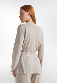 Dlouhý béžový kardigan s dlouhými rukávy a páskem na zavazování, jemná textura, detail na zadním švu a malá růžová etiketa u výstřihu.