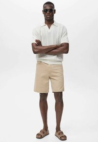Short beige shorts with a textured white short-sleeve knit polo. Accessories include dark sunglasses and brown slide sandals.