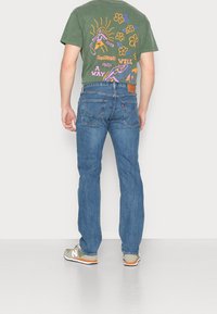 Homme portant un jean bleu, un t-shirt graphique vert avec des dessins colorés et un texte au dos, et des baskets beiges avec des touches orange.