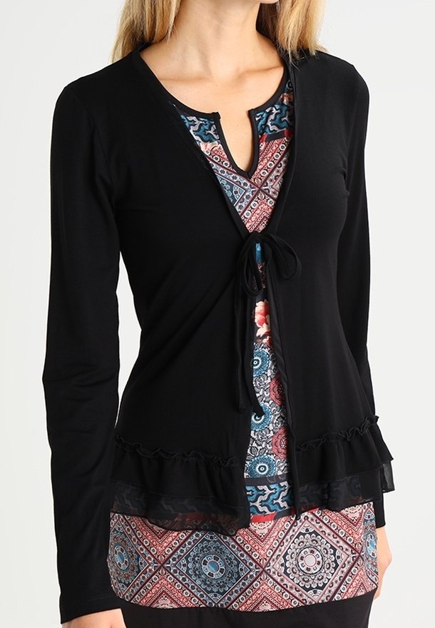 Sort langærmet cardigan med flagrende kant, bundet i midten. Båret over en flerfarvet mønstret kjole med geometriske designs.