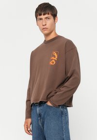 Ung mand iført en brun langærmet skjorte med en orange grafik på brystet, parret med blå denim jeans, stående mod en ensfarvet baggrund.