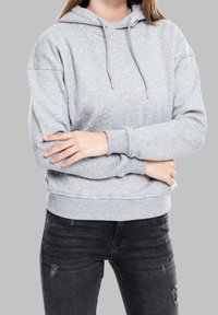 Persona vestida con una sudadera ligera gris con cordones y jeans negros desgastados, con los brazos cruzados sobre el torso.