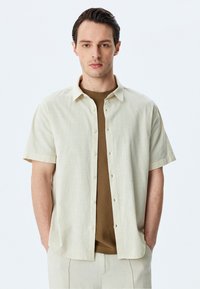 Camisa de manga corta, con botones, en tejido de lino beige claro, con una textura sutil, un cuello y un corte relajado. Llevada sobre un top de punto marrón.