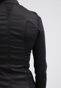 Giacca nera aderente con una texture liscia, cuciture visibili sul retro e un design strutturato che accentua le spalle.