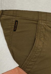 Pantalons vert olive avec une texture lisse, présentant une étiquette noire cousue sur la poche avant. La taille est blanche avec des détails côtelés.