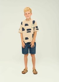 Beige T-shirt med mörkblå penseldragmönster och grön palmträdsgrafik, i kombination med denimshorts och bruna sandaler.