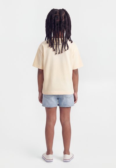 Παιδί με μακριά πλεξούδα, που στέκεται πλάτη, φοράει μπεζ μπλουζάκι, ανοιχτό μπλε τζιν σορτς και λευκά αθλητικά σε απλό φόντο.