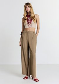 Top corto beige, pantaloni a gamba larga color sabbia, collana rossa a più strati e sandali con tacco rossi. Tessuti lisci e leggeri con una vestibilità su misura.