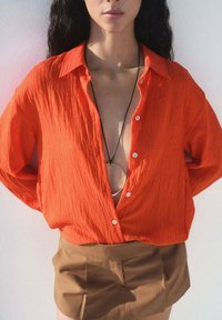 Ljust orangefärgad texturerad skjorta med djup V-ringning och knapppdetaljer, tillsammans med ett par bruna högmidjade shorts. En cirkulär halsband ingår.