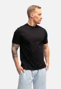 T-shirt di cotone nera con maniche corte e colletto a girocollo, abbinata a pantaloni sportivi azzurri chiari. Il modello ha un tatuaggio sul braccio destro.