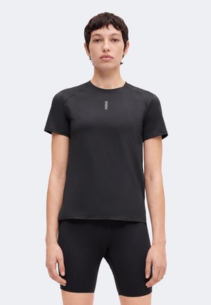 Junge Person mit kurzen dunklen Haaren, die ein schwarzes kurzärmeliges Sportshirt und schwarze enganliegende Shorts vor weißem Hintergrund trägt.