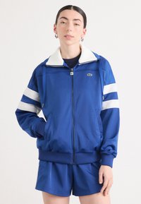 Casaco azul com fecho éclair e colarinho branco, apresentando detalhes em listras brancas nas mangas. Feito de tecido suave, inclui bolsos e punhos canelados.