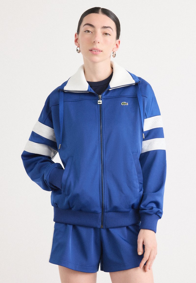 Casaco azul com fecho éclair e colarinho branco, apresentando detalhes em listras brancas nas mangas. Feito de tecido suave, inclui bolsos e punhos canelados.