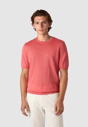 Junger Mann steht vor einfarbigem Hintergrund, trägt einen korallenfarbenen Kurzarmpullover und hellbeige Hose, schaut mit neutralem Ausdruck nach vorn.